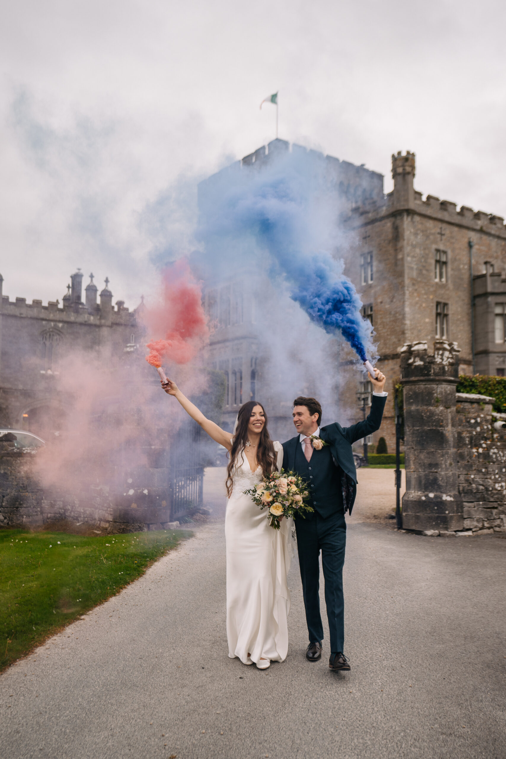 Bride and groom celebrating with red and blue smoke bombs in front of Markree Castle – joyful wedding moment full of colour and excitement.
