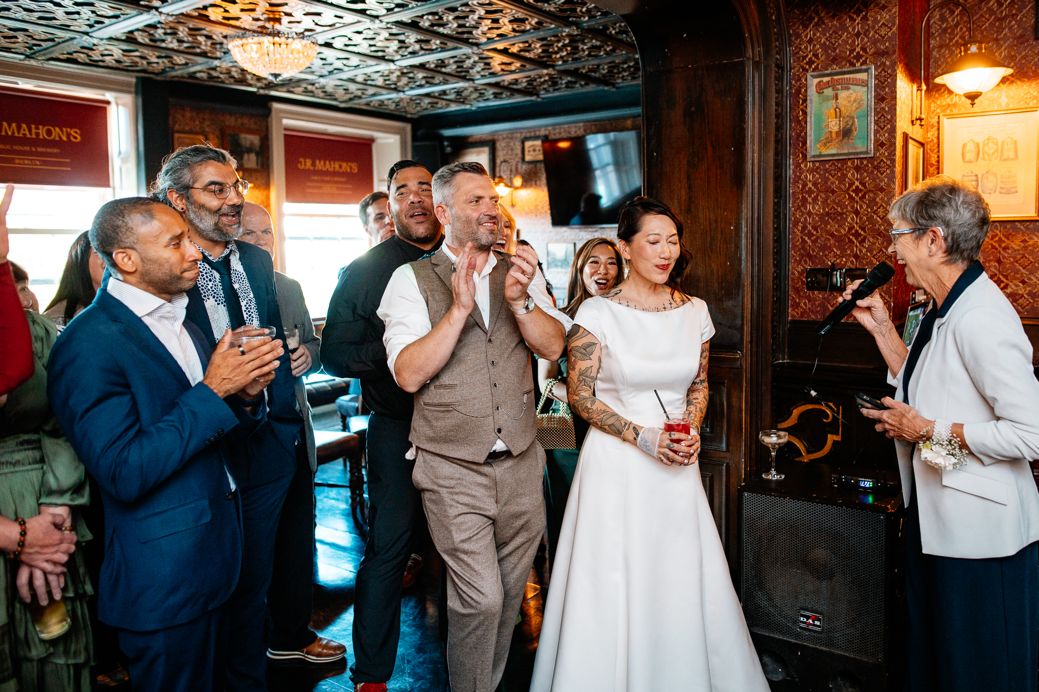 Man giving a wedding speech with a microphone as guests smile beside him at J.R. Mahon's Dublin