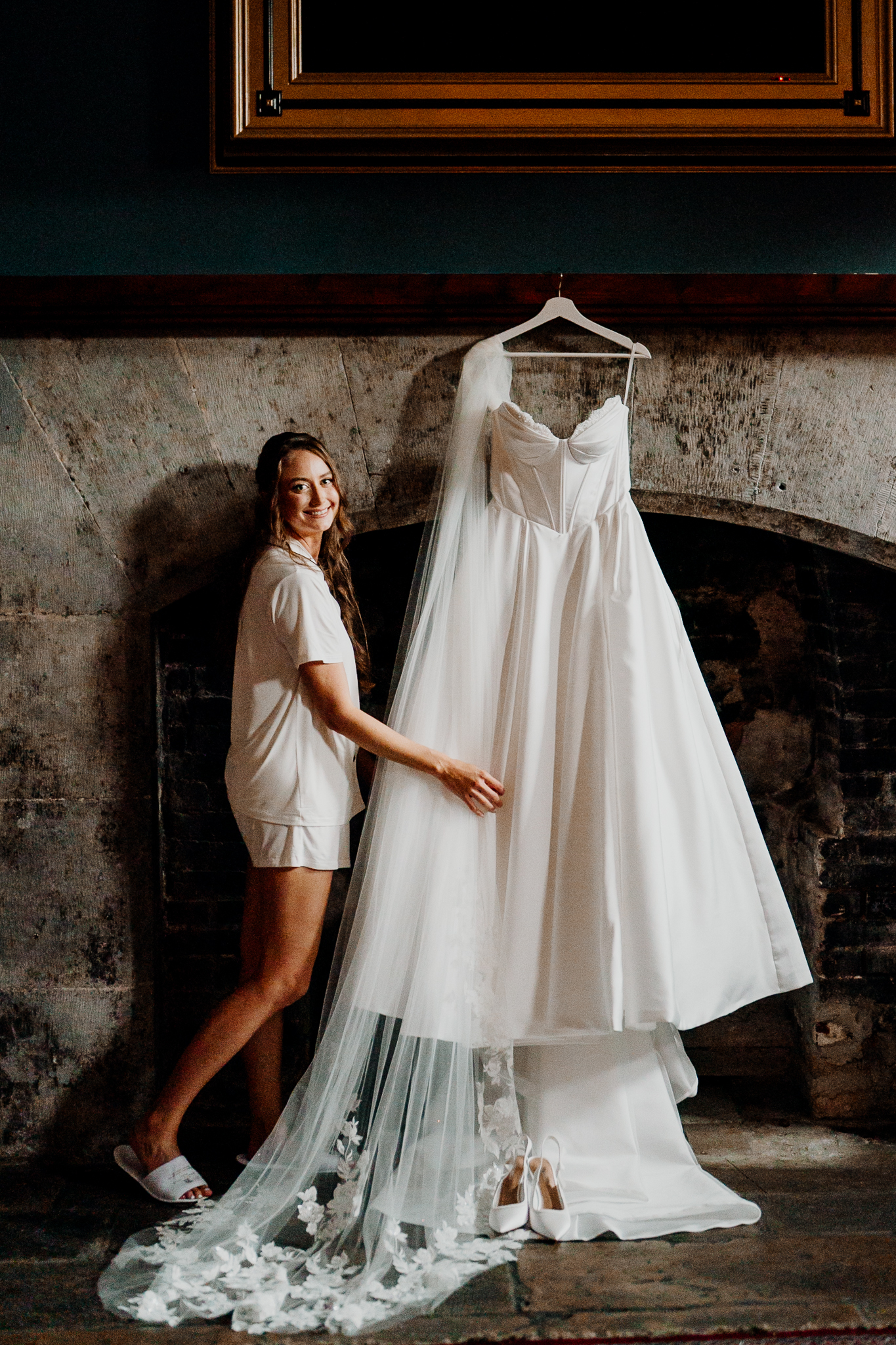Bride standing beside her hanging white wedding dress against a rustic stone wall in the Gin Bar at Markree Castle.
