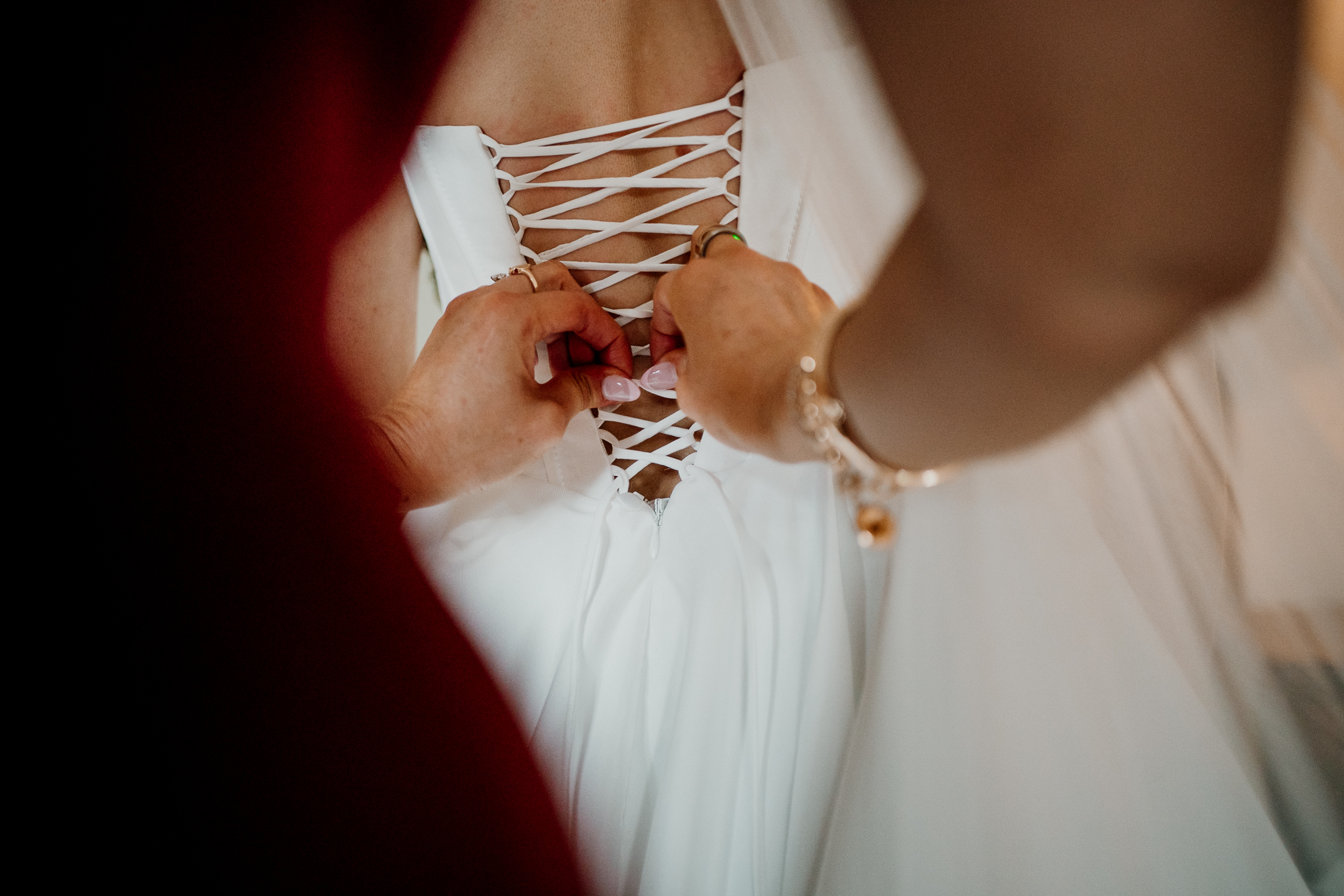 Close-up of hands fastening the lace-up corset back of a white wedding dress during bridal preparations.