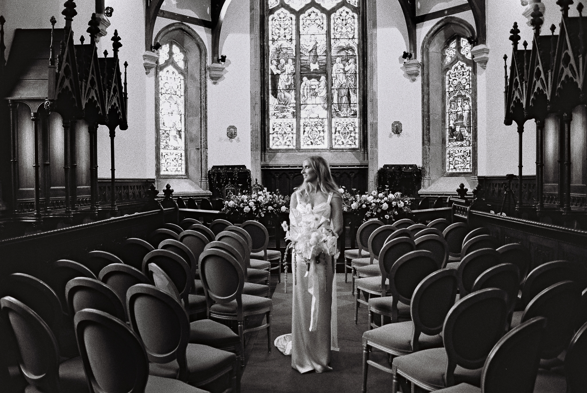 Black and white 35mm film photograph of a bride seen from behind inside Markree Castle Chapel, with historic stained-glass windows in the background.
Kopiuj
