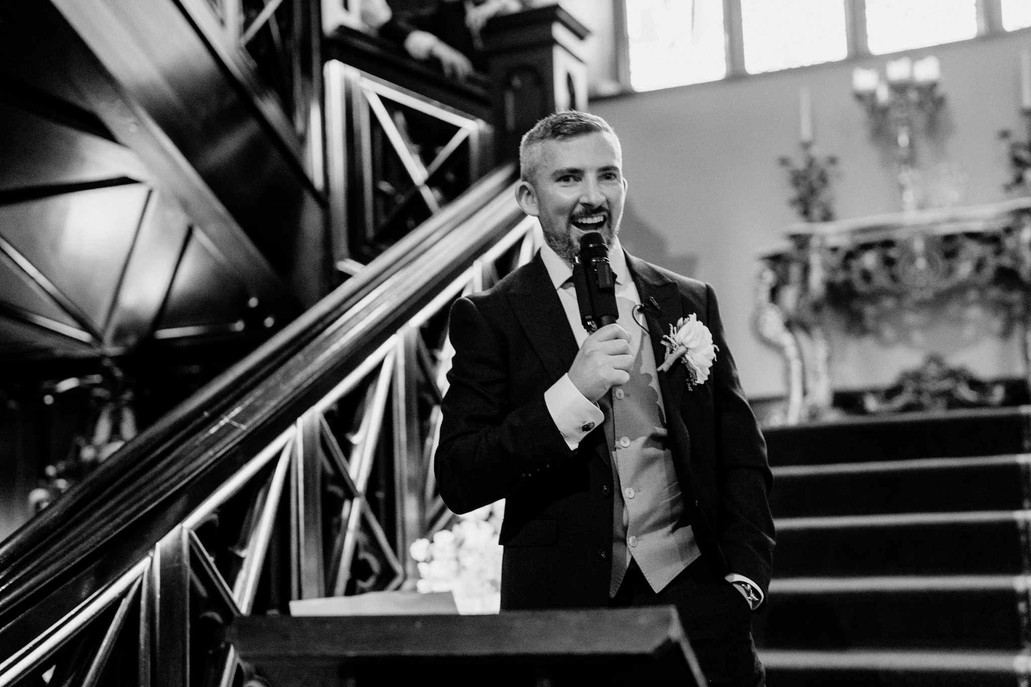 Black and white photograph of a man delivering a wedding speech with a microphone beside the grand staircase at Markree Castle.