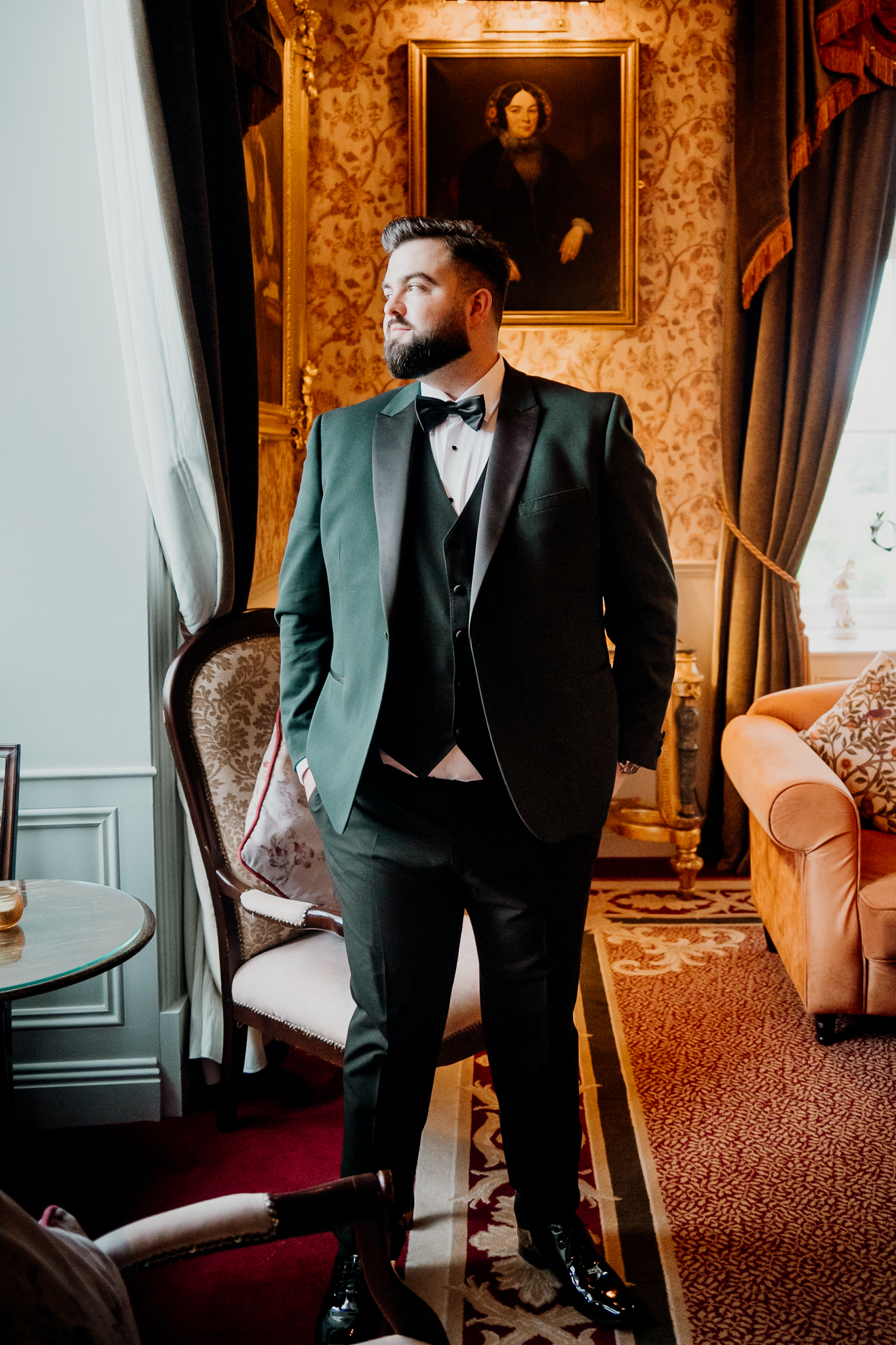 Groom wearing a dark green tuxedo and black bow tie standing beside a window in the Billiard Room at Markree Castle.