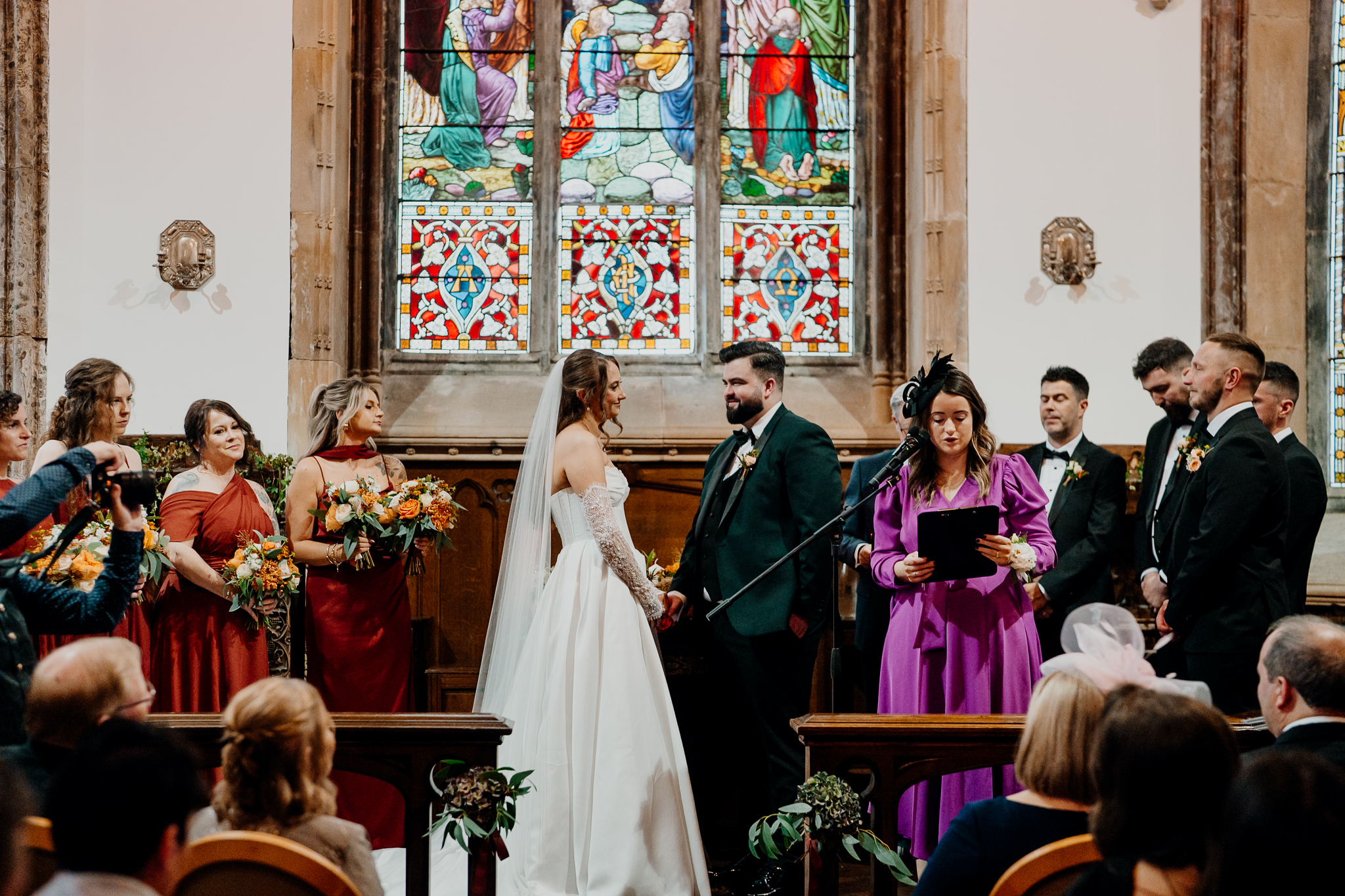 Wedding guests seated in wooden pews during an indoor ceremony at Markree Castle Chapel, beneath colourful stained glass windows.