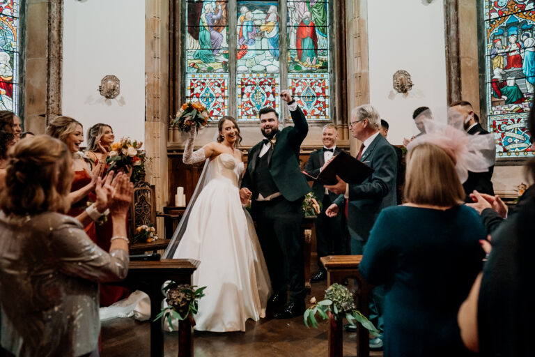 Newly married couple smiling and celebrating with guests inside Markree Castle Chapel beneath colourful stained glass windows.