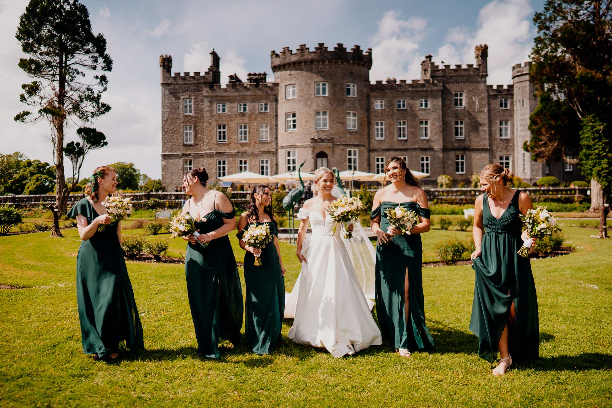 Bride walking with bridesmaids in dark green dresses carrying floral bouquets in front of Markree Castle.