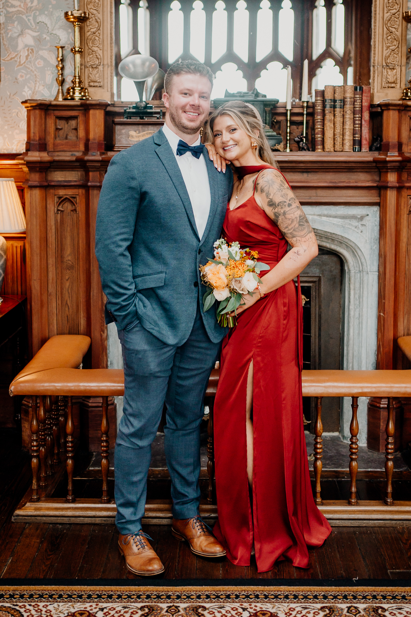 Wedding guest in a blue suit posing with a smiling woman holding an autumn bouquet beside a fireplace at Markree Castle.